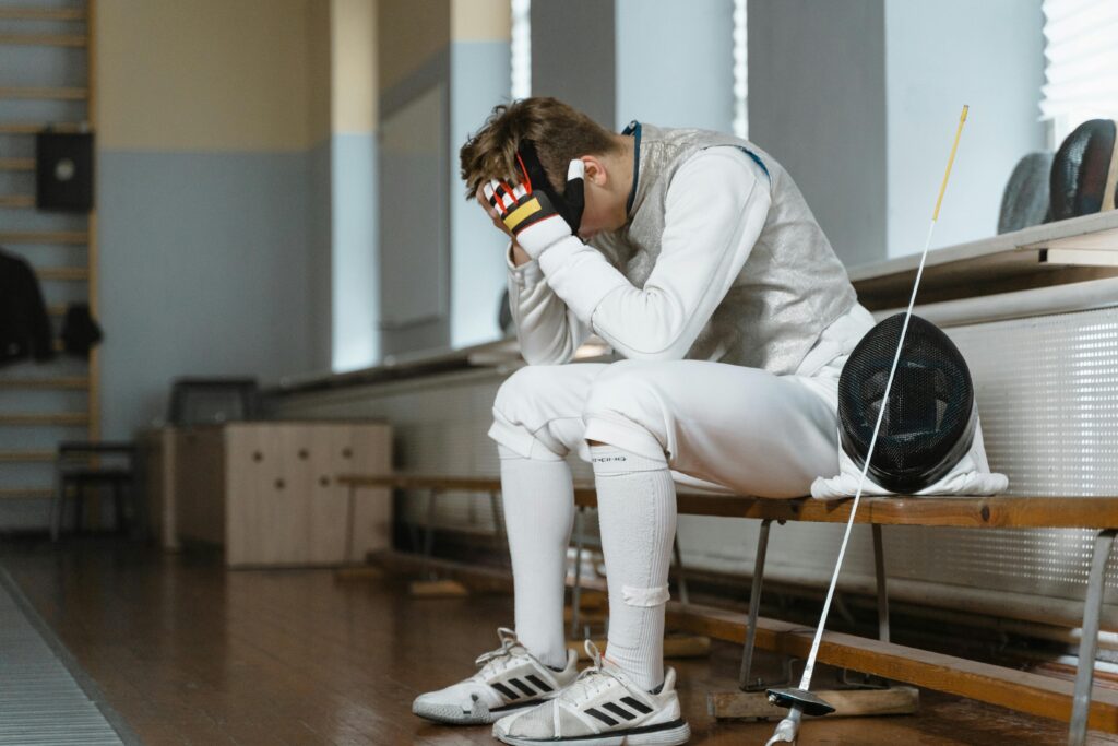 pexels-photo-6538990-6538990 Young fencer with head in hands, showing frustration during indoor training session.